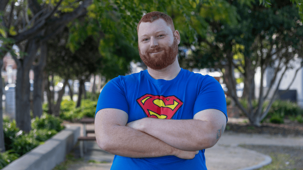 A red-haired man with a beard wearing a blue Superman T-shirt stands smiling with his arms crossed in a park.