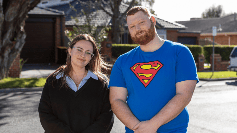 A young woman wearing glasses and a red-haired man in a blue Superman T-shirt stand side by side on a suburban street.
