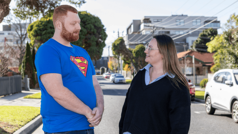 A young woman and a red-haired man stand smiling at each other on a suburban street.