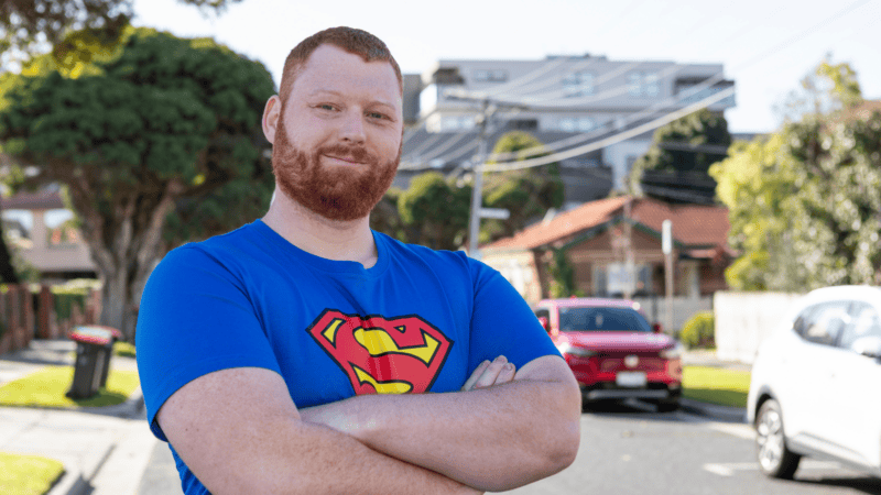 A red-haired man with a beard wearing a blue Superman T-shirt stands smiling with his arms crossed on a suburban street.
