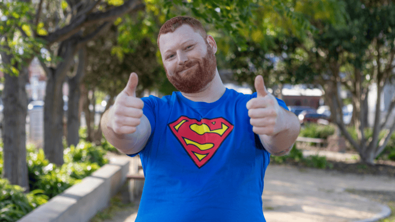 A smiling red-haired man wearing a blue Superman T-shirt gives two thumbs up outdoors.