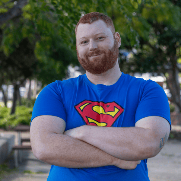A red-haired man with a beard wearing a blue Superman T-shirt stands smiling with his arms crossed in a park.